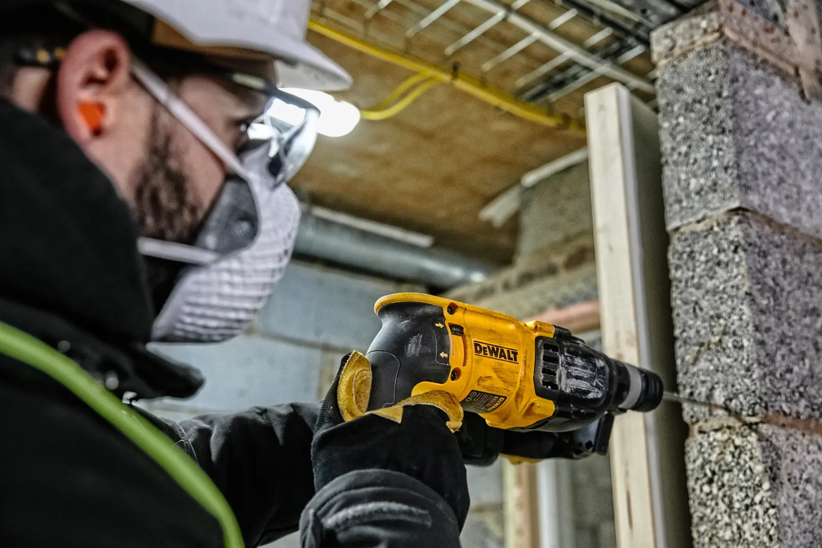 Ein Arbeiter mit Sicherheitsausrüstung verwendet einen gelben DEWALT-Bohrhammer (Modell D25133K), um eine Betonwand auf einer Baustelle zu bohren.