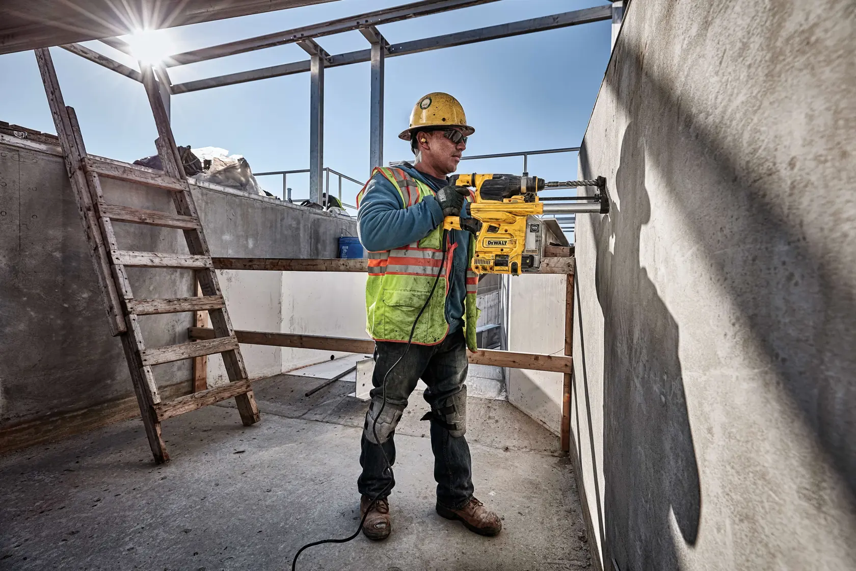 Ein Bauarbeiter mit Schutzhelm und reflektierender Weste verwendet ein DEWALT D25333K Elektrowerkzeug, um eine Betonwand auf einer Baustelle zu bohren.