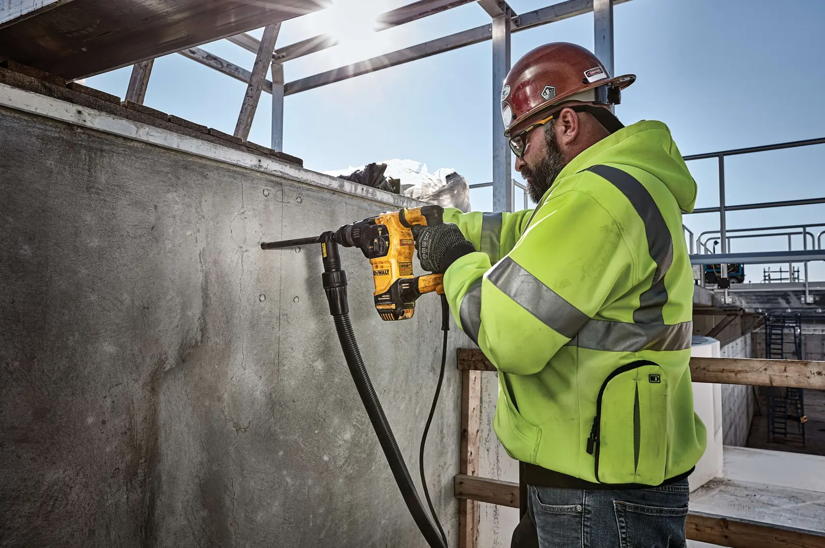 Person mit Warnjacke und Schutzhelm benutzt einen DEWALT-Bohrhammer, um eine Betonwand auf einer Baustelle zu bohren.