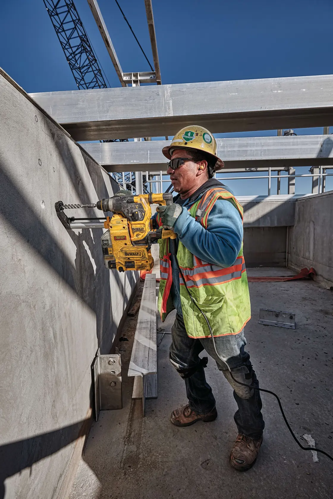 Ein Bauarbeiter mit Sicherheitsweste, Helm und Handschuhen bedient einen DEWALT-Bohrhammer (Modell D25333K), um eine Betonwand auf einer Baustelle im Freien zu bohren, umgeben von Metallträgern und Bauelementen. Das Gesicht des Arbeiters ist absichtlich verschwommen.