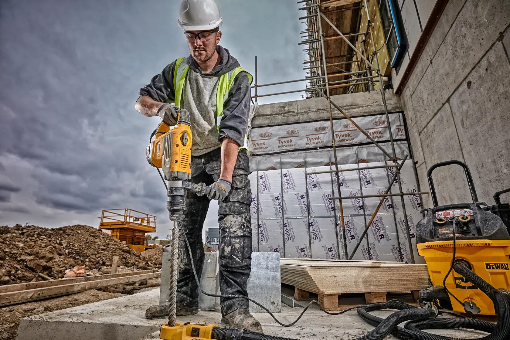 Ein Bauarbeiter mit Sicherheitshelm und Warnweste arbeitet mit einem DEWALT-Rotationshammer (SKU: D25733K) auf einer Baustelle. Das Gesicht des Arbeiters ist verschwommen, und im Hintergrund sind Baumaschinen, Gerüste und Isoliermaterialien zu sehen.