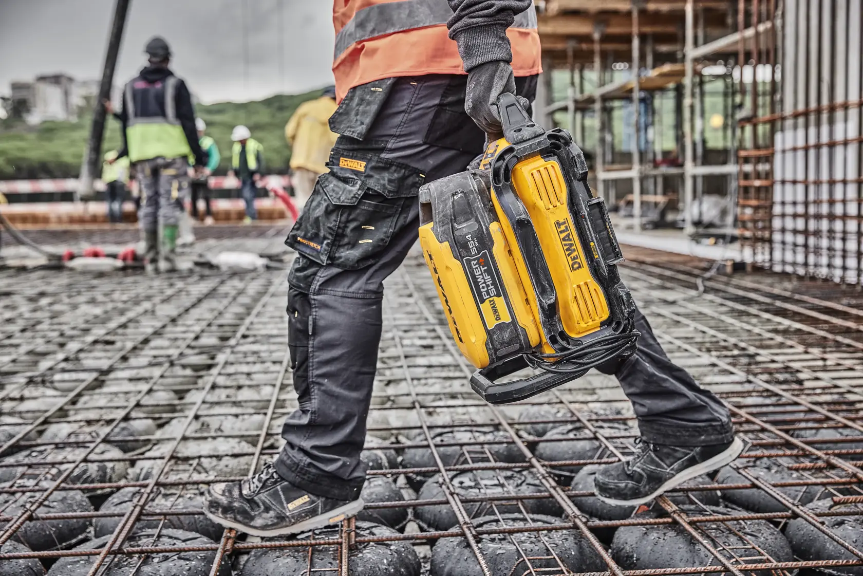 Person hält DEWALT DCBPS0554 Akku auf Baustelle, trägt Handschuhe und Jeans.