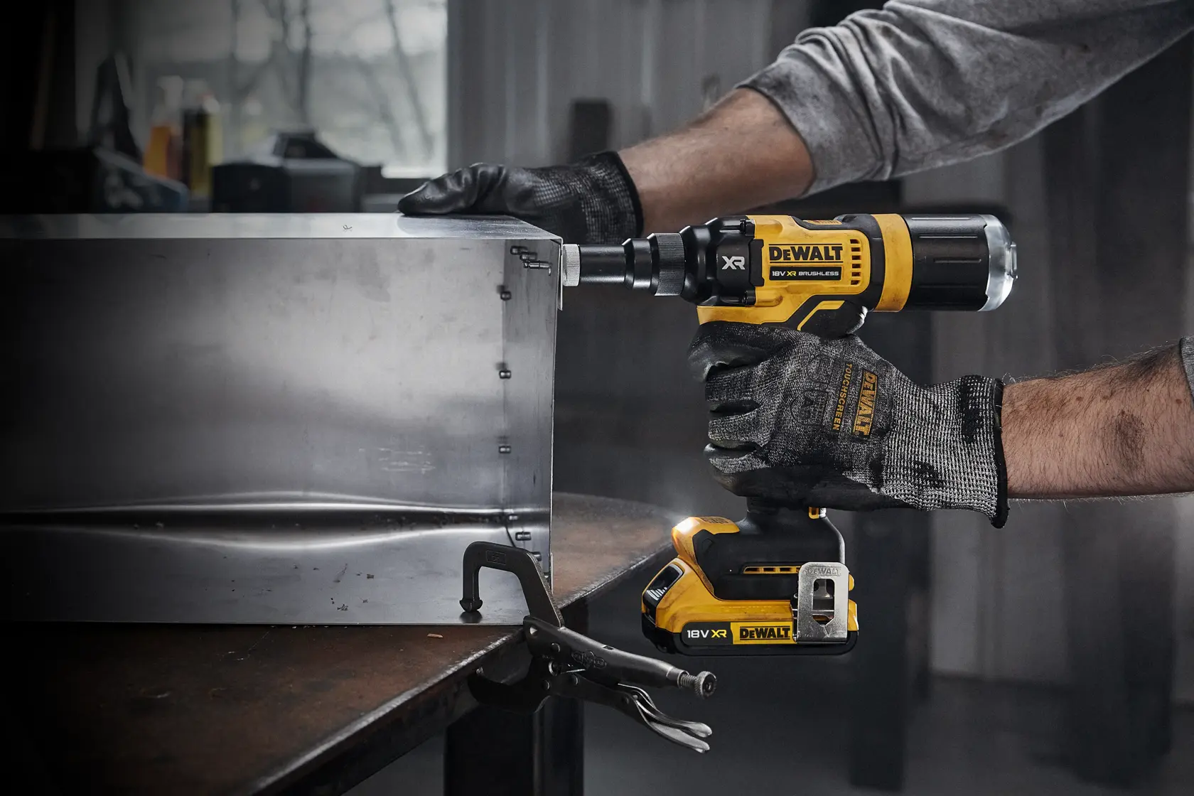 A person wearing work gloves is using a DEWALT DCF403NT cordless power tool to fasten screws into a metal box in a workshop.