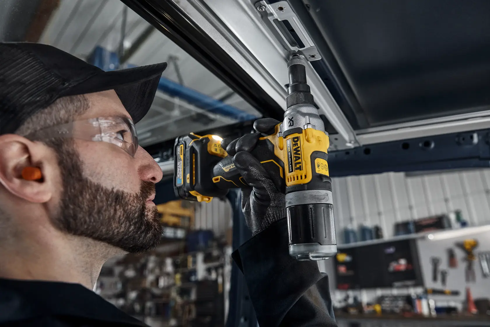 A person wearing ear protection and gloves uses a DEWALT DCF414 cordless tool to fasten or drill into a metal frame, with a workshop visible in the background.