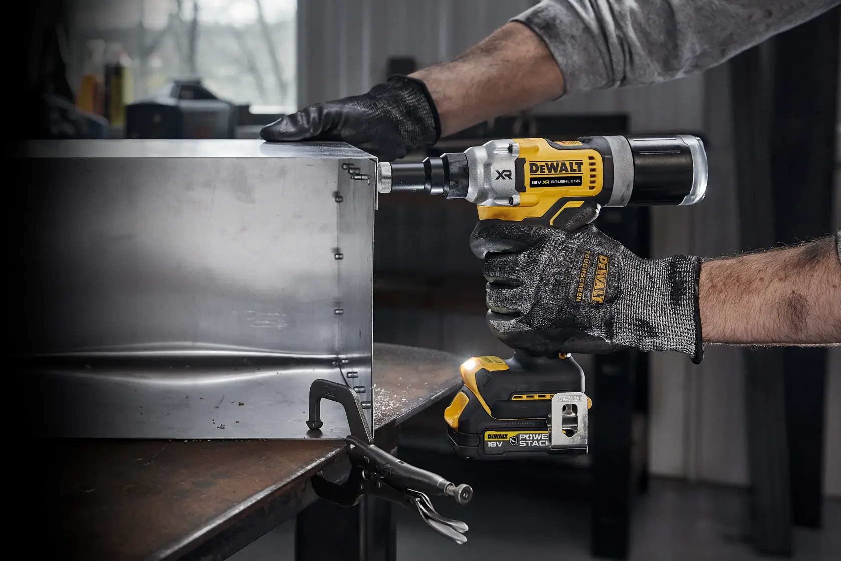 A person wearing gloves is using a DEWALT 18V XR cordless rivet tool (model DCF414) to fasten metal sheets on a worktable in a workshop.