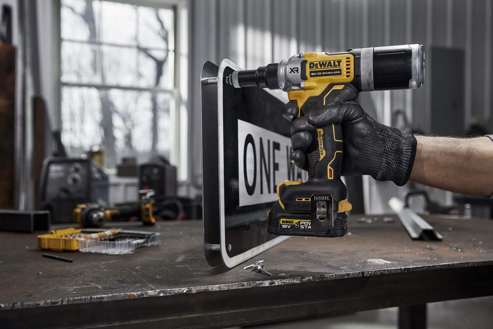 A person wearing a black glove uses a DEWALT DCF414NT cordless power tool on a metal sign in a workshop. The tool features yellow and black DEWALT branding and is positioned against the edge of the sign. Various tools and parts are visible on the workbench in the background.