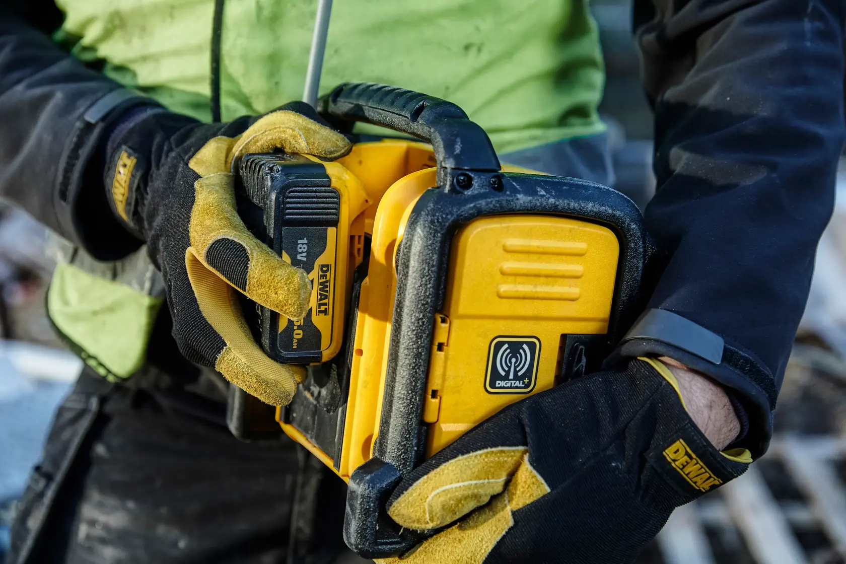 Person holds DEWALT DCR020 yellow digital radio with 18V battery, wearing work gloves at construction site.