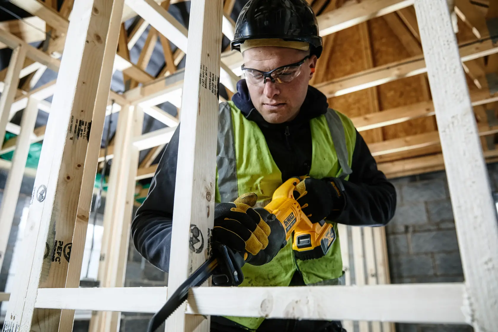 Person using a DEWALT reciprocating saw to cut wood in a house under construction, wearing safety gear.
