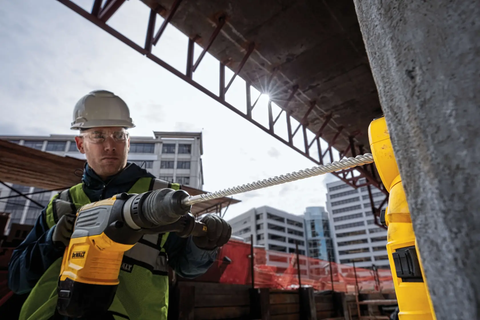 Arbeiter nutzt einen DEWALT-Bohrer auf einer Baustelle, Gebäude im Hintergrund.