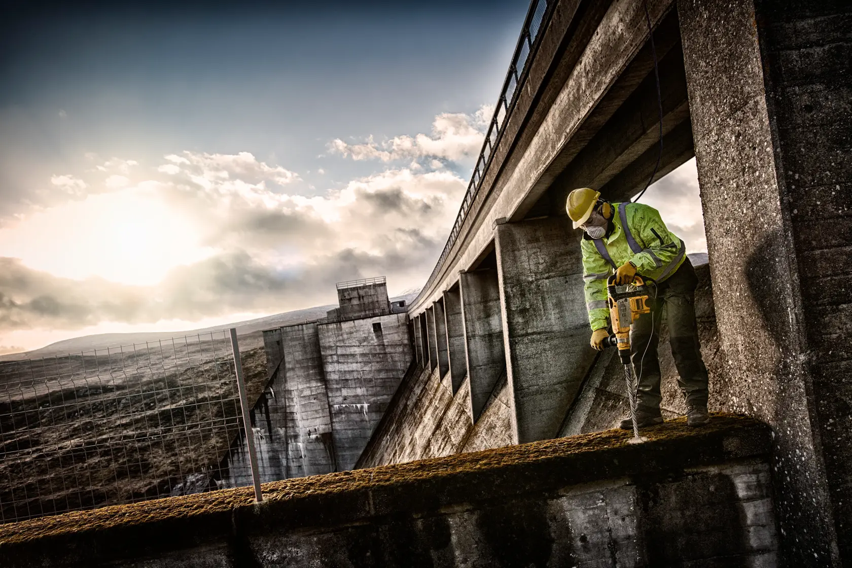 Arbeiter nutzt DEWALT-Bohrmaschine auf Beton, trägt Schutzkleidung im Freien unter bewölktem Himmel.
