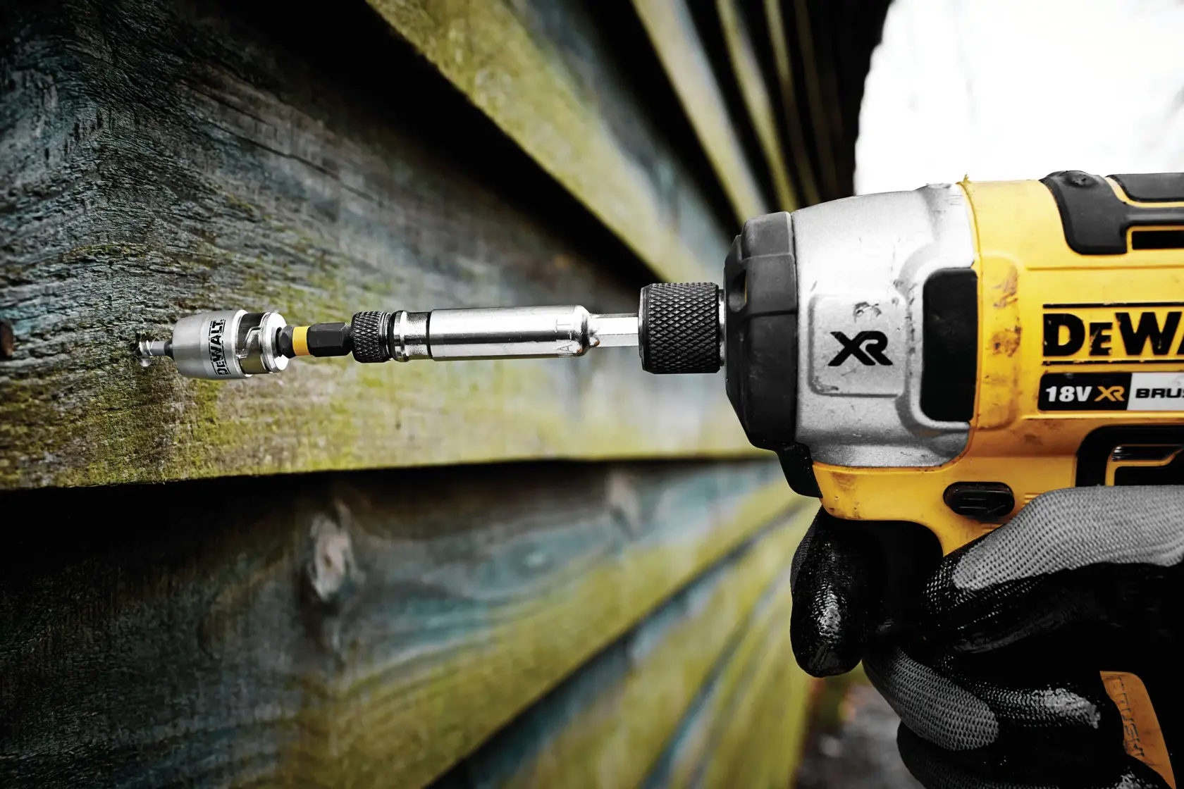 DEWALT drill with bit attached, in use on wooden siding outdoors, held by a gloved hand.