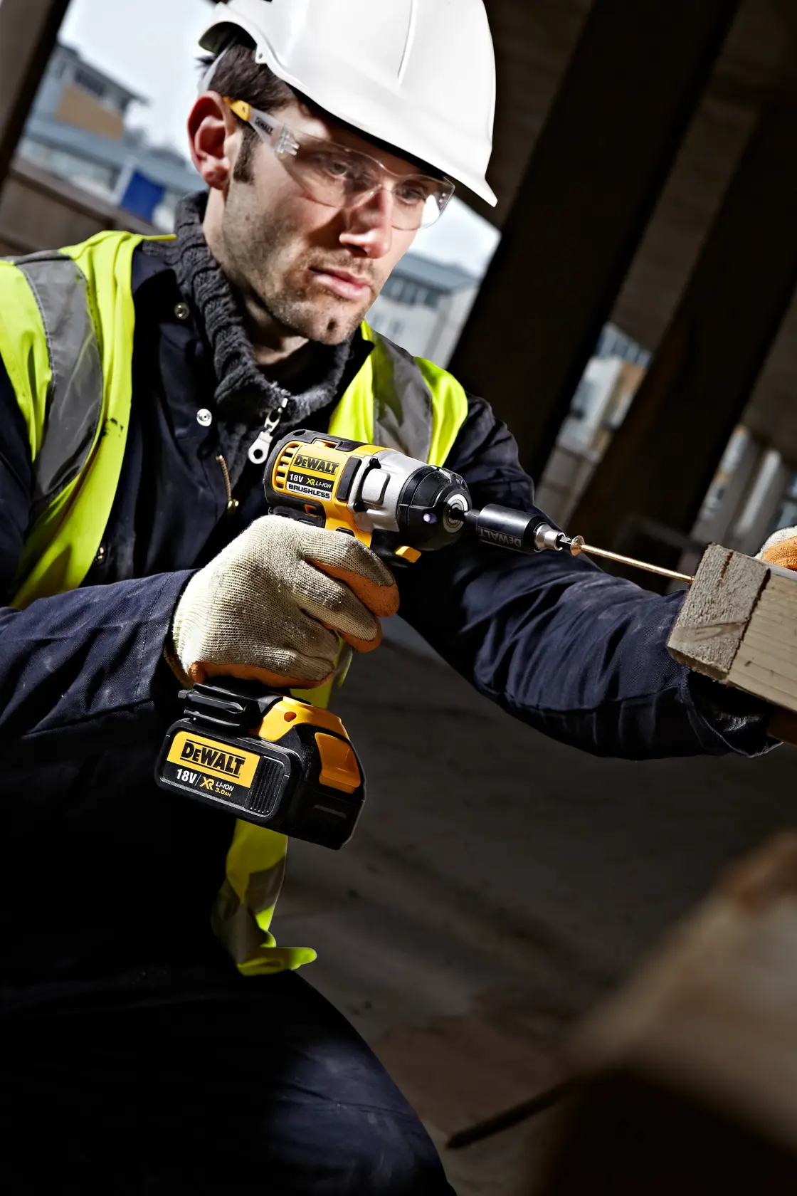 Worker uses a DEWALT cordless drill to drive a screw into wood at a construction site, wearing safety gear.