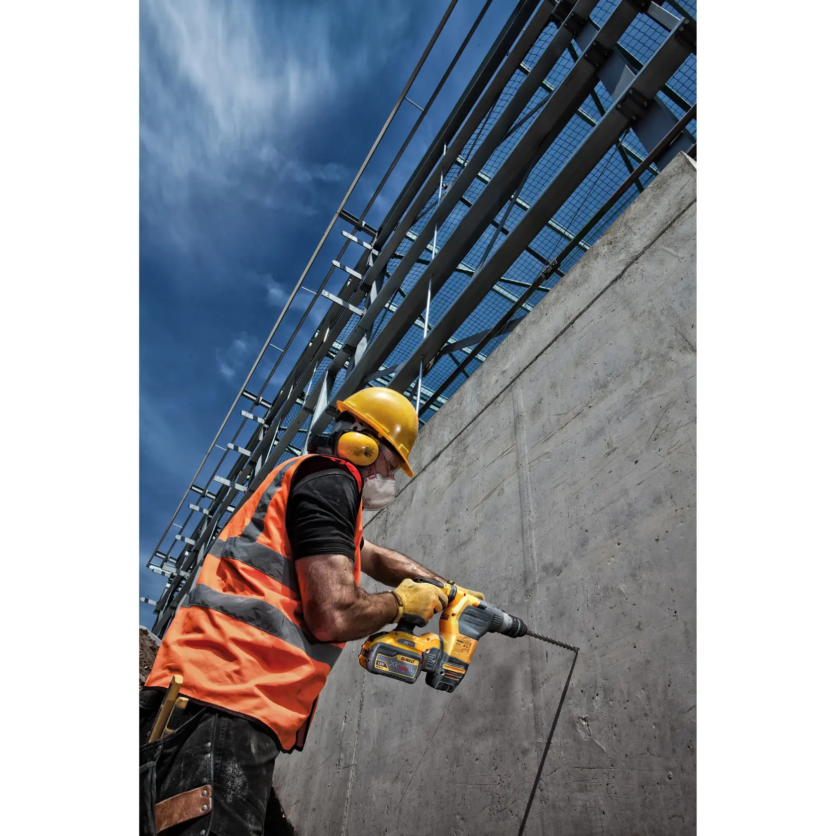 Trabajador con equipo de seguridad usa taladro DEWALT en pared de concreto en obra exterior bajo cielo azul.