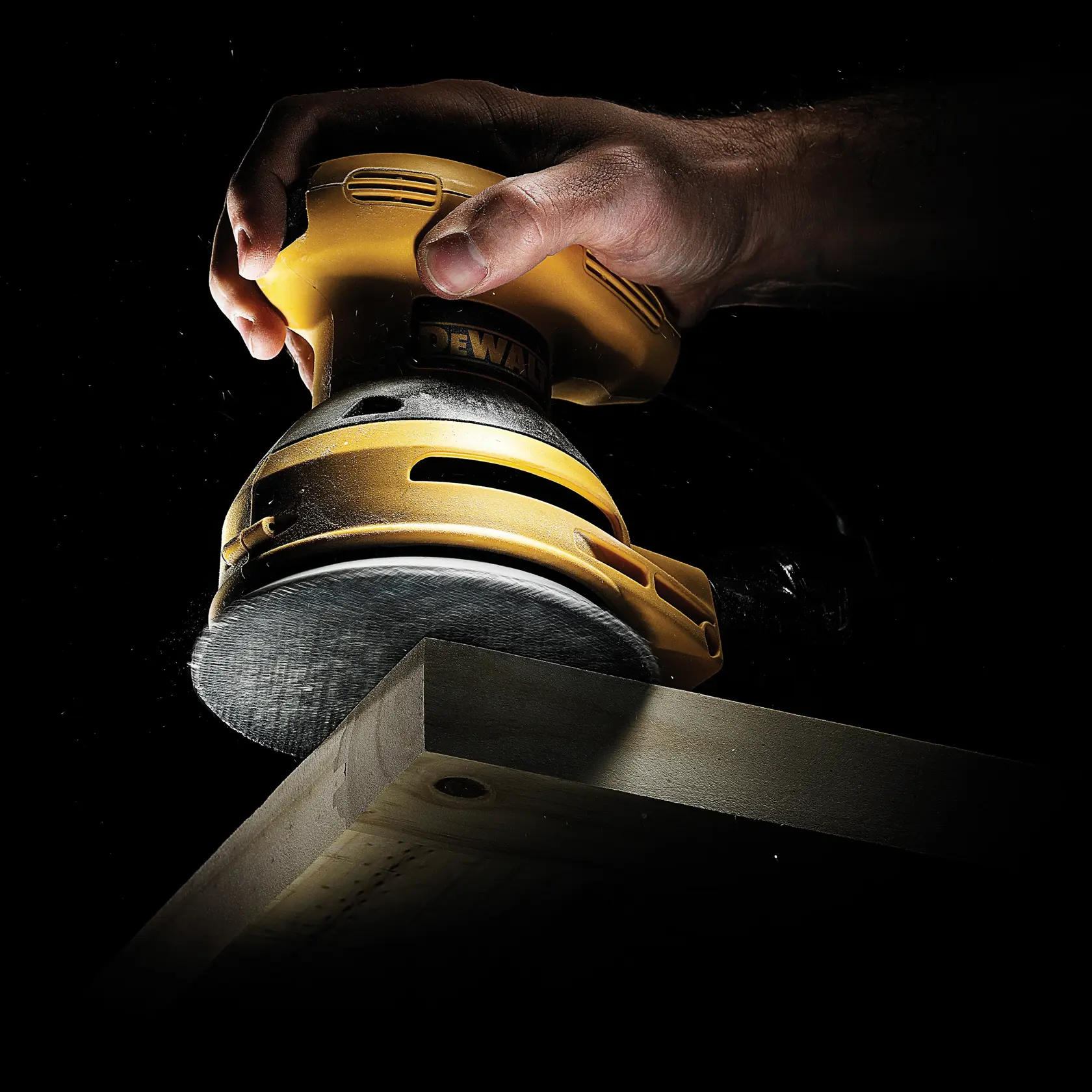 Hand using a yellow DEWALT sander to smooth the edge of a wooden plank against a black background.