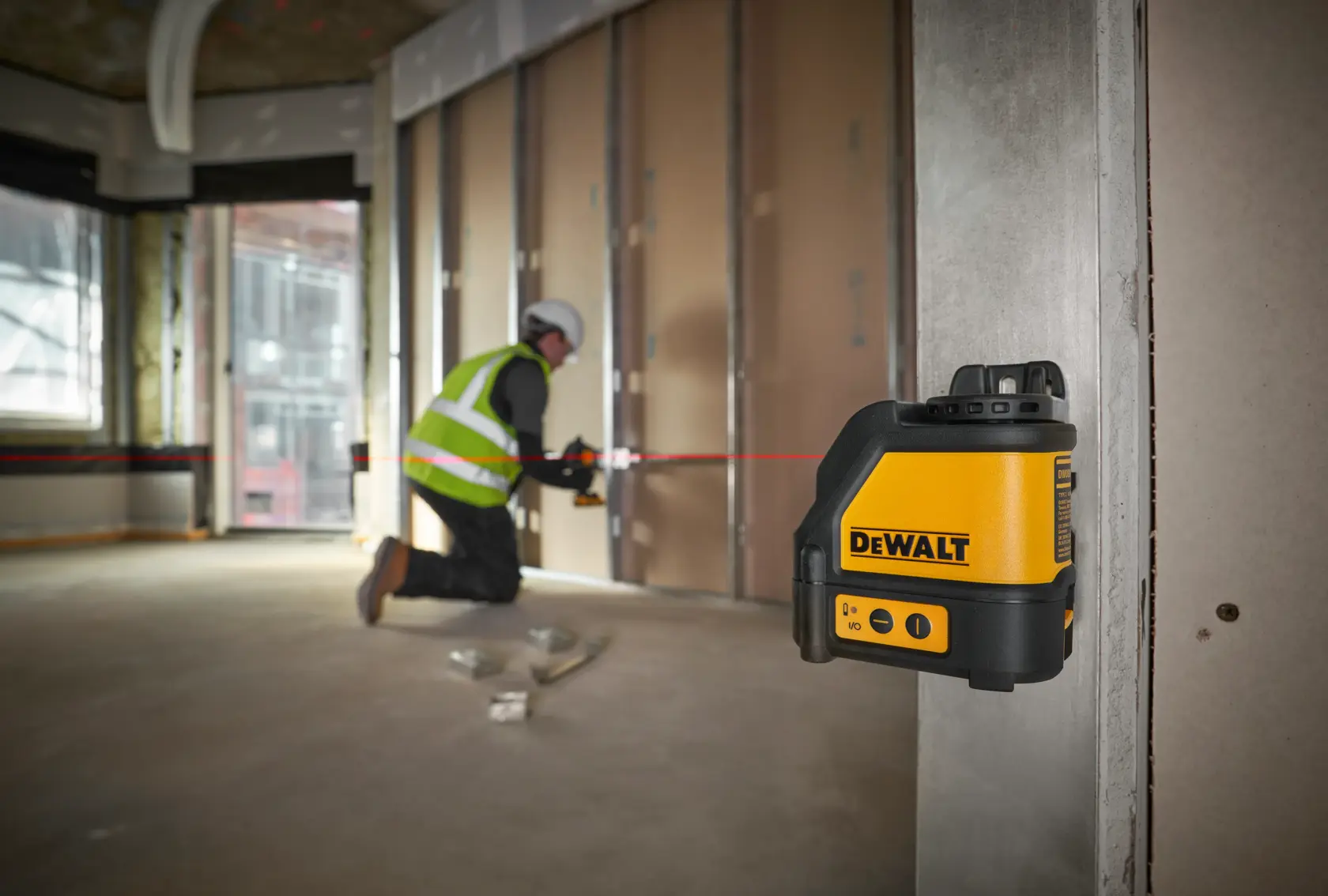 Nivel láser DEWALT DW088K proyecta línea roja en obra, trabajador instala paneles.