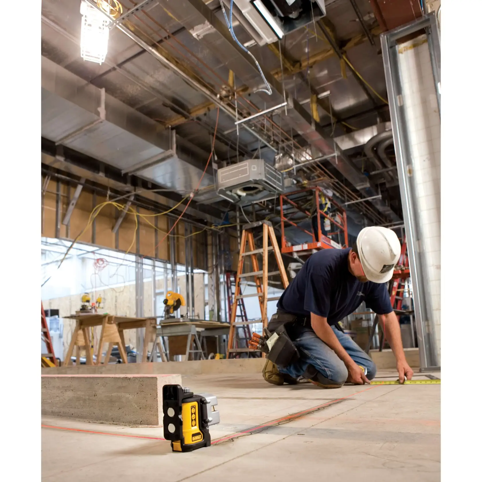Two construction workers are installing metal framing in an indoor building site. One worker stands on a ladder, while the other is kneeling on a scaffold. Both are wearing safety helmets and tool belts. A DEWALT laser level (DW089K) is placed on the floor, projecting visible red lines onto the concrete for precise alignment.