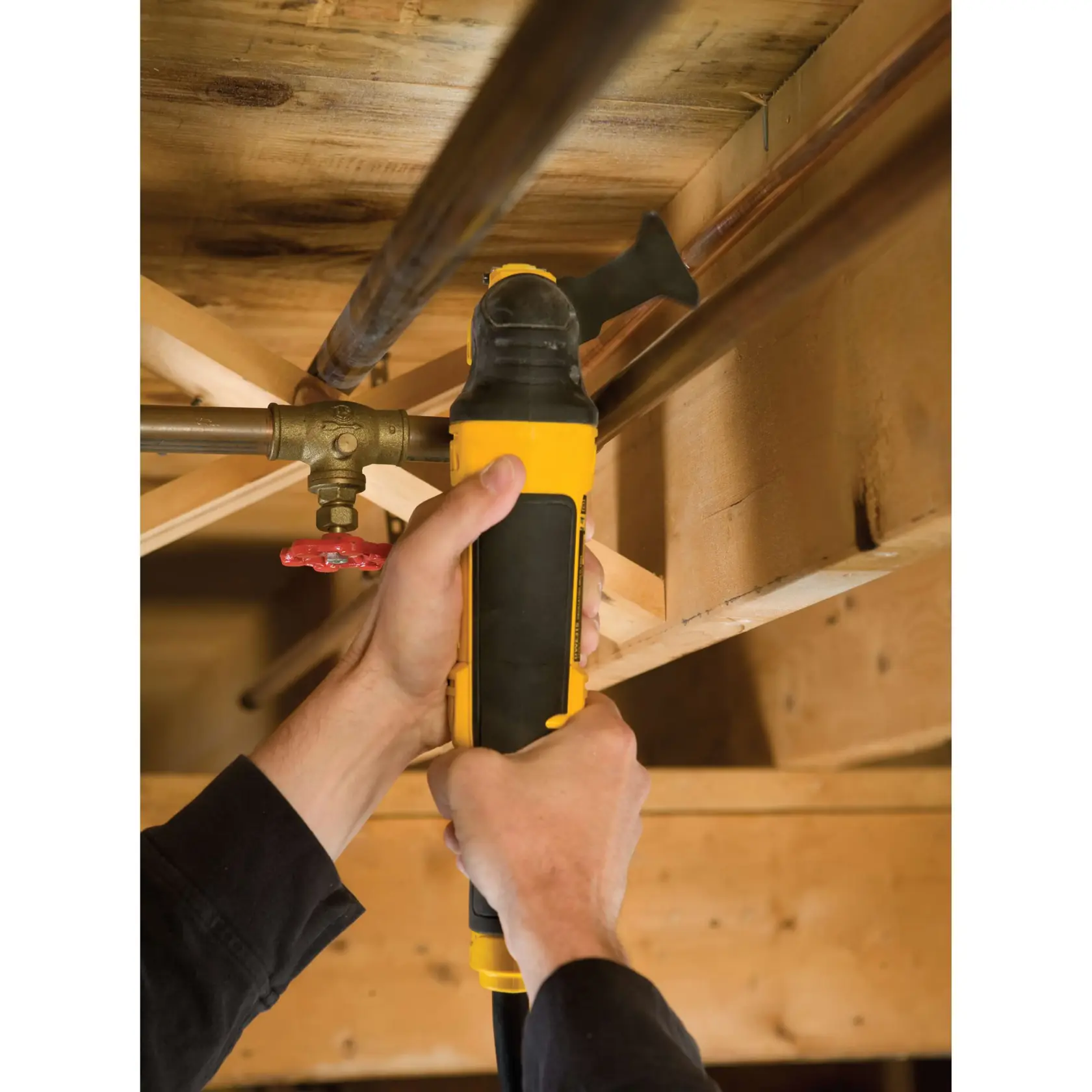 Hands using a DEWALT multi-tool on a pipe under wooden beams. Tool is yellow and black.