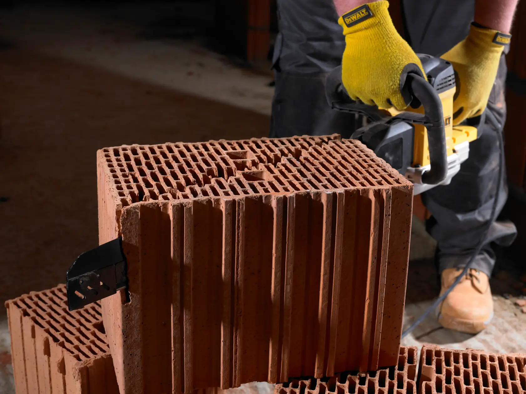 A person wearing yellow gloves is using a DEWALT power tool to cut through a large hollow clay brick. The tool is positioned against the brick and the person is standing on a dusty surface.
