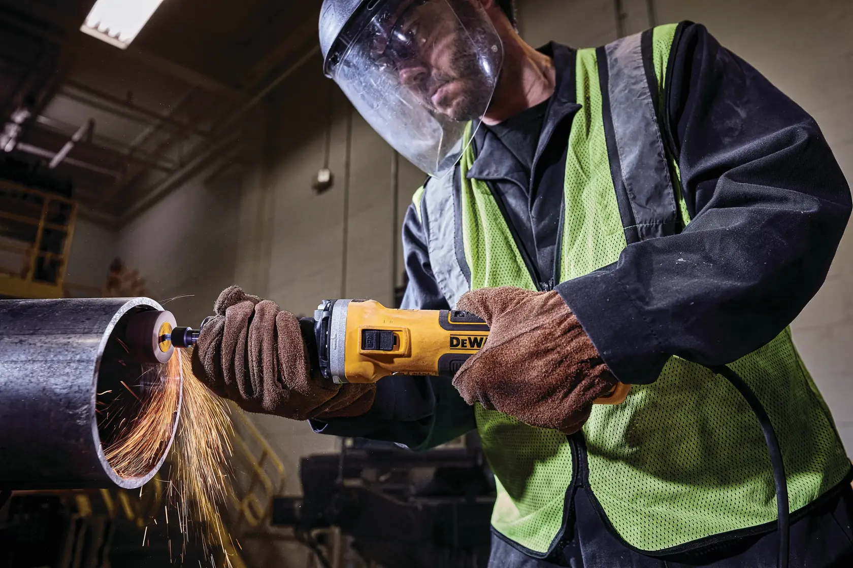 Worker using a DEWALT grinder on a metal pipe, wearing gloves, safety vest, and face shield with sparks flying.