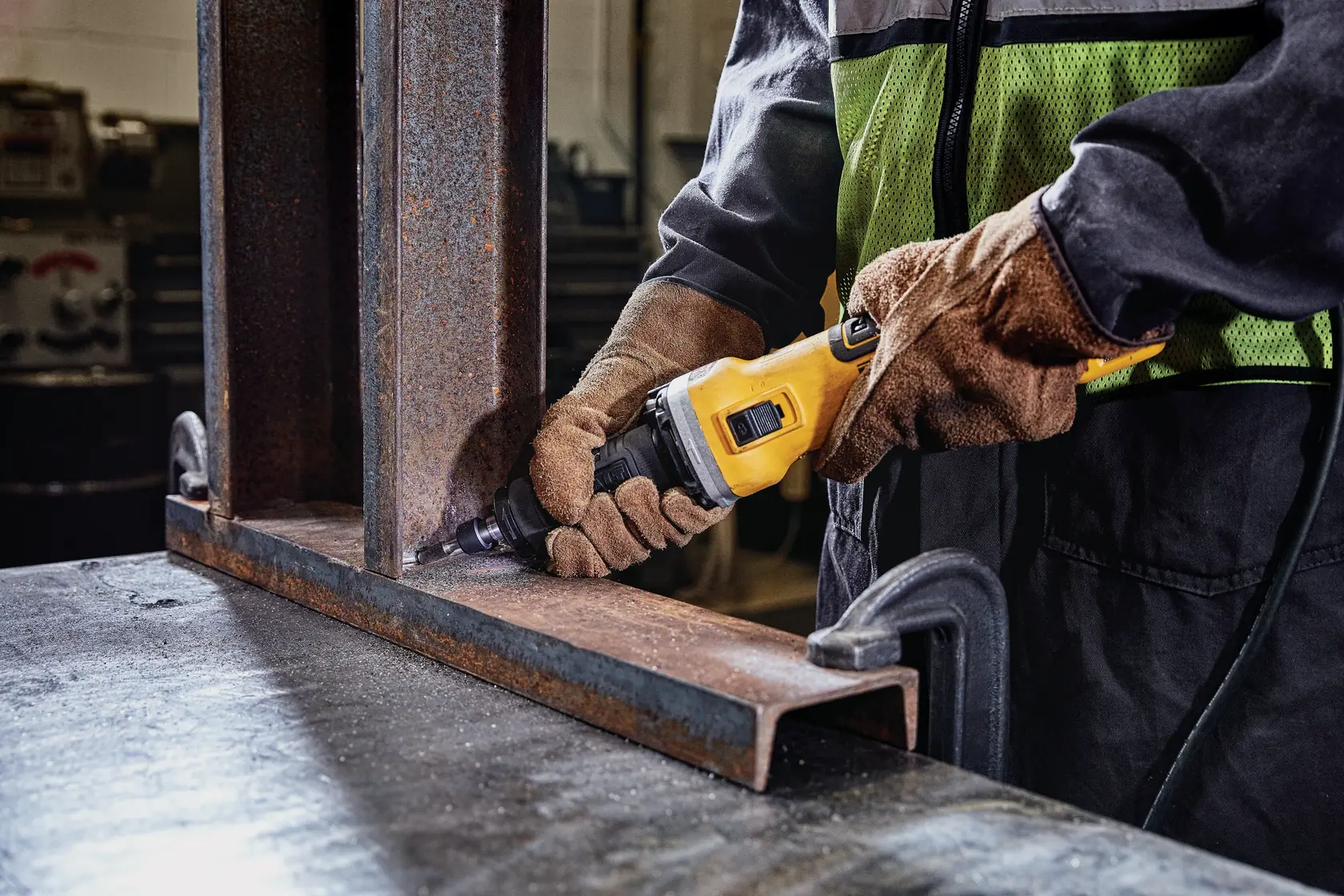 Photo of worker using DEWALT DWE4997 grinder on rusted metal beam indoors, wearing gloves and safety vest.