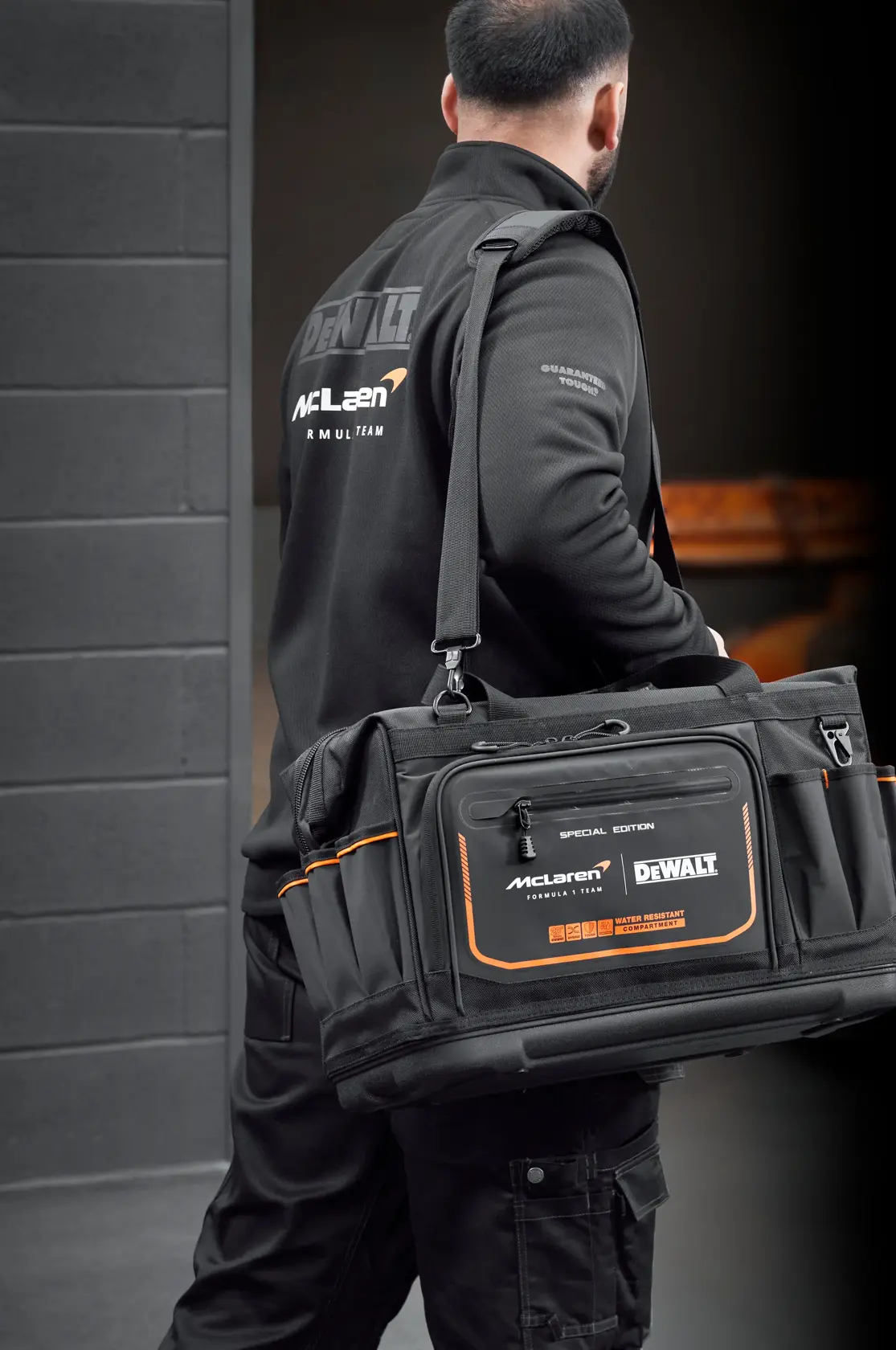 Persona lleva una bolsa de herramientas negra DEWALT McLaren con detalles naranjas en ambiente industrial.